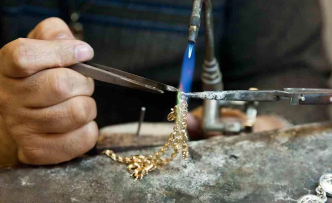 Người thợ đang hàn một sợi dây chuyền vàng bằng đèn hàn trên bàn làm việc, sử dụng kẹp để giữ chặt.