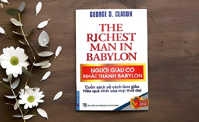 Sách "The Richest Man in Babylon" đặt trên bàn gỗ, bên cạnh là nhánh cây với hoa trắng.