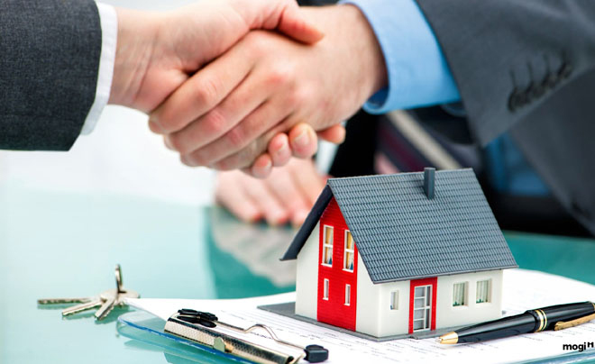 Hai người bắt tay trên bàn với mô hình ngôi nhà, chìa khóa và tài liệu, tượng trưng cho giao dịch bất động sản.