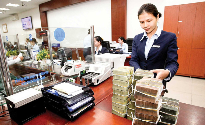 Nhân viên ngân hàng đếm tiền mặt tại quầy giao dịch, với nhiều bó tiền xếp chồng trên bàn làm việc.