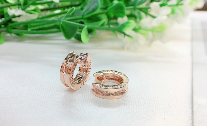 Một đôi bông tai vàng hồng được đặt trên bề mặt trắng, phía sau là nền xanh của cây cỏ.