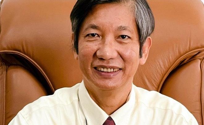 Người đàn ông trung niên mỉm cười, mặc áo sơ mi trắng, cà vạt đỏ, ngồi trên ghế da màu nâu.