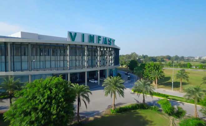 Tòa nhà lớn với tên thương hiệu trên mái, bao quanh là cây cối và con đường, dưới bầu trời trong xanh.